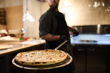 Elektrikli fırında pişirilmiş iştah açıcı İtalyan pizzasıyla metalik tepsiye odaklan ve mutfağın ortasında duran genç aşçı tarafından tut.