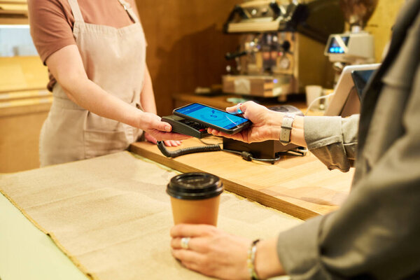 Close-up of hands of young consumer with smartphone and bakery clerk with terminal during process of contactless payment over counter
