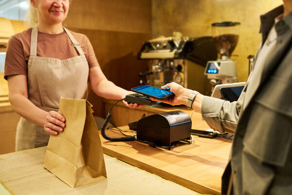Cropped shot of mature bakery clerk holding paper bag with takeaway food or pastry items and payment terminal while client paying for purchase