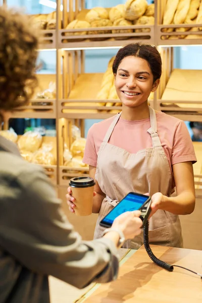購入のための非接触支払いのプロセス中に笑顔でパン屋の顧客を見てコーヒーのカップと幸せな若い女性事務員 — ストック写真