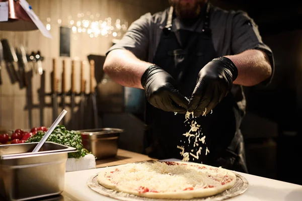 厨师们穿着黑色防护手套 站在餐馆厨房的餐桌旁 在比萨饼面包上洒上烤奶酪的特写镜头 — 图库照片