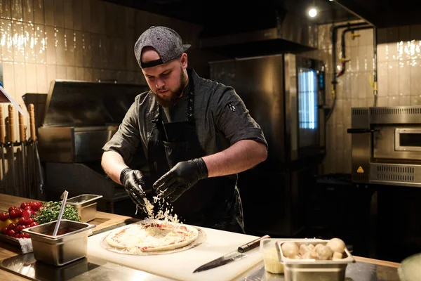 年轻厨师身穿制服 戴着黑色防护手套 在豪华餐厅厨房里做披萨时 把烤奶酪撒在扁平面包上 — 图库照片