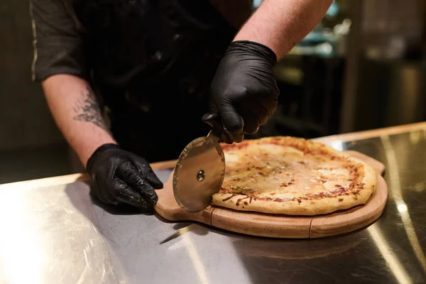 年轻厨师站在厨房桌子边 手戴黑色橡胶手套 用锋利的刀叉在木板上切开胃烤披萨 — 图库照片