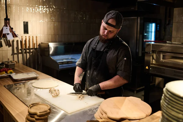 身穿黑色手套和围裙的年轻厨师在厨房里用餐桌准备披萨时 在切菜板上切碎新鲜蘑菇 — 图库照片
