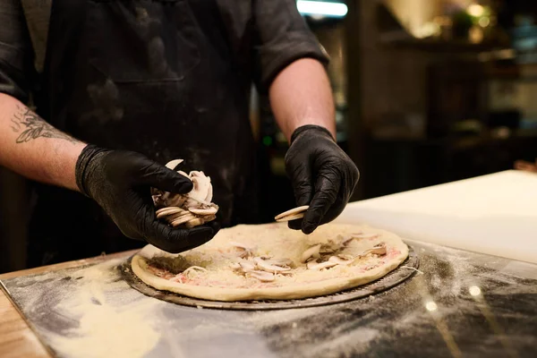 年轻厨师在厨房餐桌边做饭时 将新鲜切碎的蘑菇放在意大利披萨的扁平面包上的特写镜头 — 图库照片