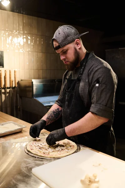 Üniformalı ve eldivenli genç sakallı adamın misafirler için pizza hazırlarken taze mantar koyuşunun yan görüntüsü.