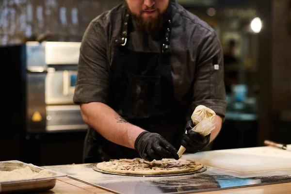 ピザを調理しながら フラットブレッド上のペストリーバッグから食材の混合物を絞る若いシェフの黒い手袋で手 — ストック写真
