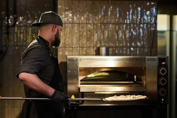 身穿工作服 留着胡子的年轻人站在厨房的热烤箱前 手里拿着圆圆的铁盘或铁架上的披萨 — 图库照片