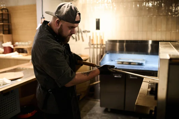 年轻厨师站在餐厅厨房用金属夹子从热电炉中取出烤比萨饼的侧视图 — 图库照片