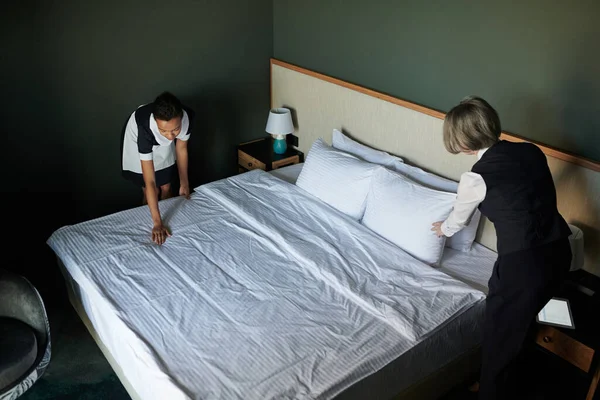 新しいゲストのための均一な準備室内で若い客室メイドを支援するホテルのヘッドマネージャー一方 両方のダブルベッドの上に曲げ — ストック写真