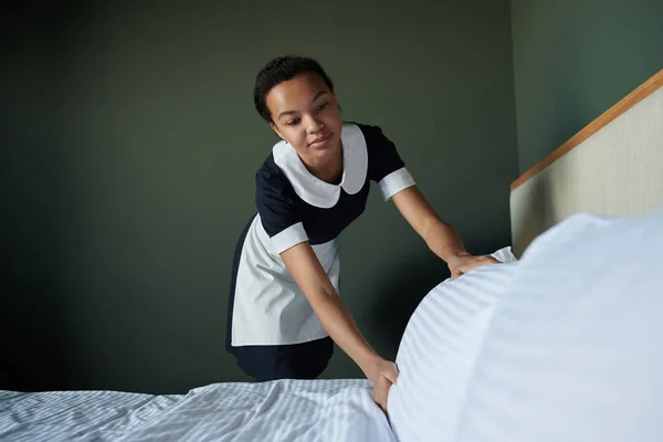 若いアフリカ系アメリカ人の部屋のメイドで制服を着た新しいゲストのためのダブルベッドを準備しながらベッドの服を変更し 枕を矯正 — ストック写真