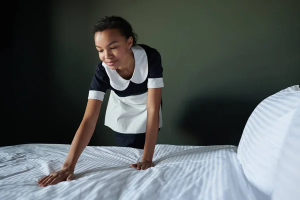 年轻的非洲裔美国女服务员弯腰坐在舒适的双人床上 一边换毛毯 一边为客人准备床上用品 — 图库照片