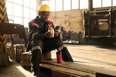 Aç genç mühendis yemek molasında ahşap kutuda oturup sandviç yerken haberlere ya da akıllı telefondaki paylaşımlara bakıyor.