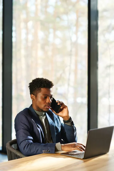 年轻而严肃的非裔美国经纪人坐在笔记本电脑前靠窗的位置上 一边用智能手机讲话 一边打字 — 图库照片