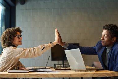 Başarılı genç erkek ve bayan meslektaşlar iş yerinde dizüstü bilgisayarlar, boncuklar ve kağıt belgelerle birbirlerine beşlik çakıyorlar.