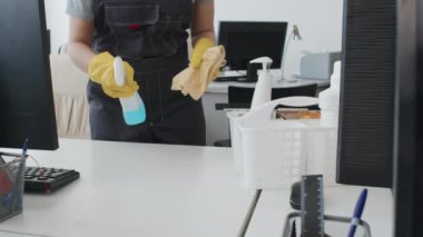 Orta bölüm, üniforma giyen, sarı lastik eldivenli, ofiste masaları deterjan ve kumaşla temizleyen bir kadın.
