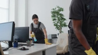 Modern ofiste masa silme ve elektrikli süpürge kullanma gibi etnik çeşitlilikte genç kadın ve erkeğin ağır çekimde çalışması.