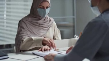 Tesettürlü ve koruyucu maskeli genç Müslüman bir kadın danışma sonrası panoya tıbbi belge imzalıyor ve her ikisi de masada otururken doktorun elini sıkıyor.