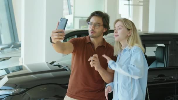 ブログのための新しい車を購入することについてのスマートフォンの録画ビデオを保持ディーラーショールームに立つ現代の若いカップル — ストック動画