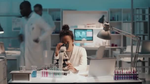 顕微鏡で化学物質を研究し ノートを作成し コンピュータを使用し 職場でアフリカ系アメリカ人男性の同僚と対話する研究室のコートで忙しい若い多民族女性の経過 — ストック動画