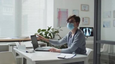 Koruyucu maskeli genç bayan doktor dizüstü bilgisayar katlıyor, tesettürlü Müslüman hastayı klinik ofisine davet ediyor ve hastanede tıbbi danışmanlık sırasında onunla konuşuyor.