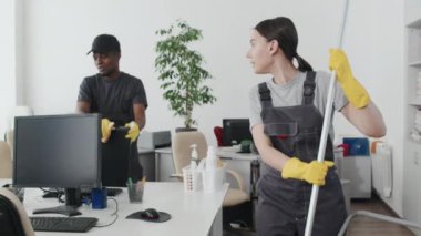 Etnik çeşitliliğe sahip genç ve neşeli bir adam ve kadının ofiste yerleri paspaslaması ve bilgisayar monitörlerini silmesi.