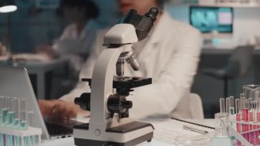 Dişi bilim adamının mikroskopta incelediği, bilgisayarını kullanan ve çalışan belgeye not alan yakın çekim zamanı. Laboratuvarda çalıştığı yerde mataralarda sıvı maddeleri incelerken.