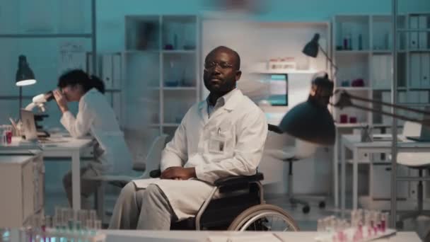 在化学实验室的研究工作中 非洲裔美国残疾男性科学家坐在轮椅上 看着摄像机与一群来回走动的实习生对视的时间 — 图库视频影像