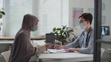 Üniformalı ve koruyucu maskeli genç tıp personeli, kliniklerde istişareden sonra imzalamak üzere tesettürdeki Müslüman kadın hastaya tıbbi belge dağıtıyorlar.