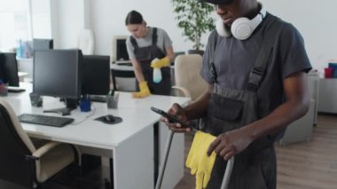 Seçici odak, yavaş çekim, temizlik işinde çalışan Afro-Amerikalı genç adamın orta ölçekli görüntüsü, iş yerindeki haberciyi kontrol ediyor.