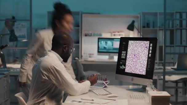 研究室のコートに身を包んだアフリカ系アメリカ人男性科学者のタイムラプスは 顕微鏡画面上で細胞のイメージを持つコンピュータの前に座り 研究を行い 同僚と協力して — ストック動画