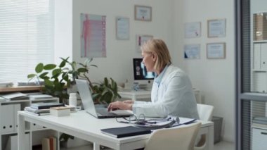 Olgun radyolog dizüstü bilgisayarın önünde işyerinde oturuyor, klavyedeki düğmeye basıyor, online hastanın akciğer röntgenini çekiyor ve uzaktan muayene sırasında görüntüye bakıyor.