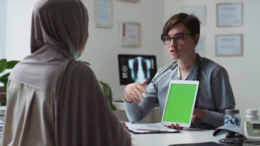 Kliniklerde tesettürde tesettüre girmiş Müslüman kadın hastaların önünde otururken elinde boş yeşil ekran tabletle üniformalı kendinden emin bir sağlık görevlisi.