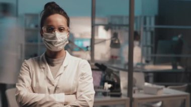 Laboratuvar önlüklü, koruyucu maskeli ve gözlüklü genç bayan laboratuvar çalışanlarının çalışma saatleri ve arka planda bir grup meşgul meslektaşıyla kameranın önünde durmaları.