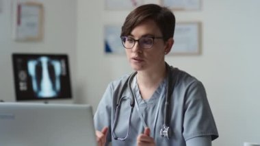 Boynunda steteskop olan mavi üniformalı genç bir kadın klinisyen bilgisayarın ve govong online danışmanlığının önünde otururken çevrimiçi hastaya yeni etkili haplar öneriyor.