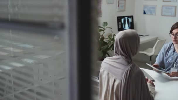 年轻自信的放射学家 身穿制服 在平板屏幕上评论脑部扫描 在演示期间坐在她面前向穆斯林女性患者解释一些细节 — 图库视频影像