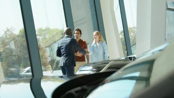 アフリカ系アメリカ人の営業担当者は ディーラーショールームで新しい車を選択するのに役立つそれらに伴う新しい顧客を迎える — ストック動画