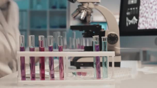 試験管内の液体を扱う防護作業服の若い女性科学者のタイムラプス閉鎖性と化学研究所の職場での顕微鏡での特性を研究する — ストック動画
