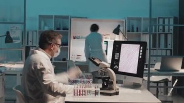 Yetişkin bilim adamlarının ve laboratuvar önlüklü, koruyucu maskeli ve eldivenli genç stajyerlerin çalışma saatleri. Klinik deneylerini masalarıyla gerçekleştiriyorlar ve sonrasında bireysel çalışmalarının sonuçlarını tartışıyorlar.