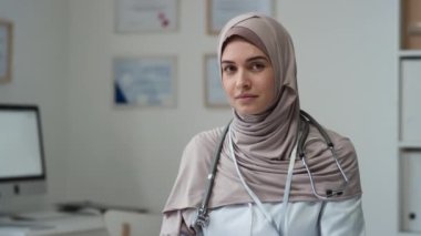 Tesettürlü, laboratuvar önlüklü, boynunda steteskop olan genç ve güzel bir bayan doktor bilgisayar monitörüne ve tıp bürosunun duvarlarında asılı sertifikalara bakarken kameraya bakıyor.