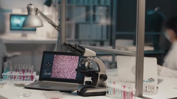 拿着手提电脑 显微镜和一组装有液体物质的试管的现代临床医生对着各自工作的身穿实验室外套和防护面具忙碌的科学家的工作时间 — 图库视频影像