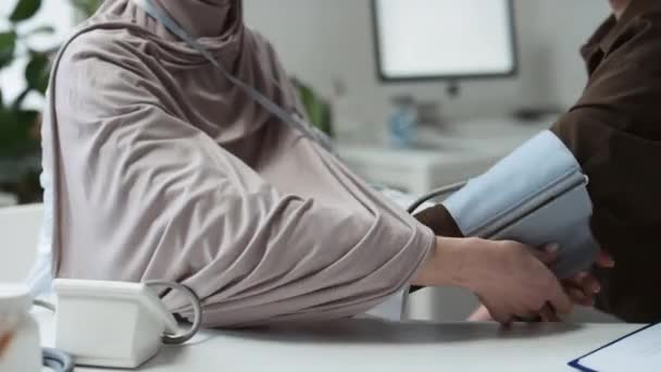 年轻的穆斯林女医生或全科医生坐在医务室临床医生的工作场所时对病人进行头巾检查血压的特写 — 图库视频影像