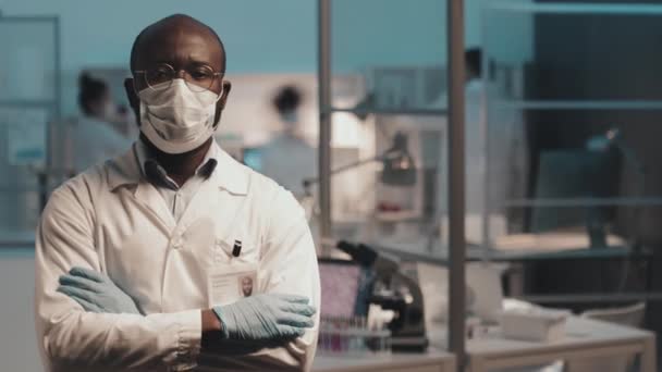身穿实验室外套 戴防护面具和戴手套的非洲裔美国男性科学家的时间在胸前交叉交叉 并对着在实验室工作的同事看着相机 — 图库视频影像