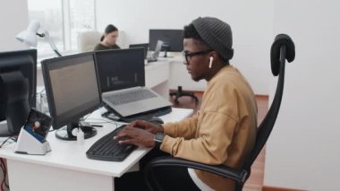 Gündelik giysiler ve kablosuz kulaklıklar giyen genç Afrikalı Amerikalı bilişim uzmanı modern ofiste kod üzerinde çalışıyor.