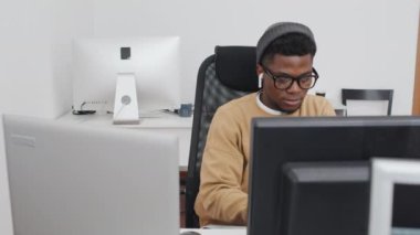 Modern ofiste masa başında oturmuş bilgisayarlarla uğraşan, gündelik kıyafetler giyen Afrikalı genç bir adamın yüksek açılı yavaşlığı.