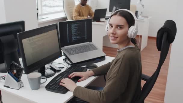 It企業のオフィスのデスクに座っているヘッドフォンを身に着けている陽気な若い白人女性プログラマーはカメラで微笑んだ — ストック動画