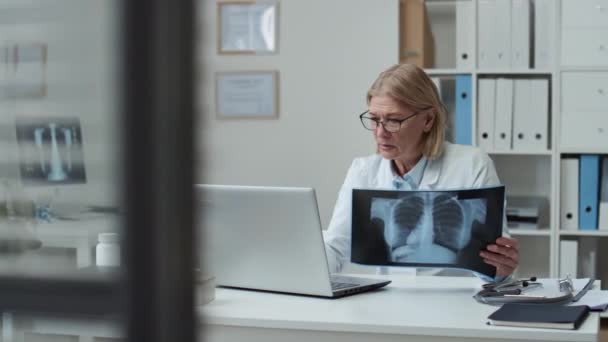 코트를 금발의 의사가 엑스레이를 노트북으로 타이핑을 병원의 진료실에서 온라인 환자와 — 비디오