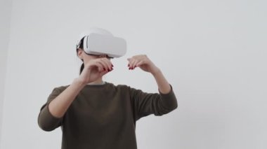 Düşük açılı orta açı çekimde beyaz kadın VR kulaklık takıyor oyun oynuyor ya da çalışıyor, beyaz duvar arkası