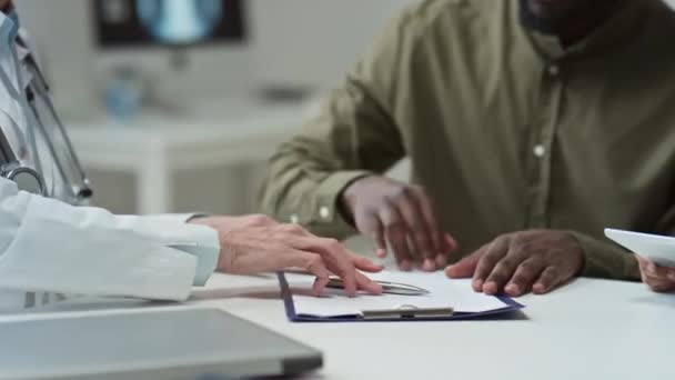 将附有医疗文件的剪贴板交给年轻的非洲裔美国男性患者 由其在工作场所握手向其签名致意的成熟女临床医生的特写镜头 — 图库视频影像