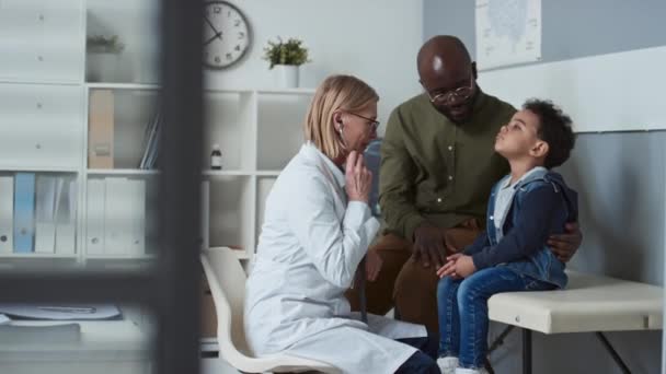 年轻的非洲裔美国男子坐在他的小儿子旁边 而成熟的女儿科医生则带着听诊器检查孩子 并在医务室与他的父亲交谈 — 图库视频影像